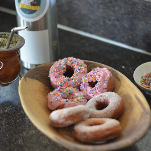 Donas sin gluten con glaseado rosa y granas de colores, y rosquillas con azúcar, en una fuente de madera junto al mate